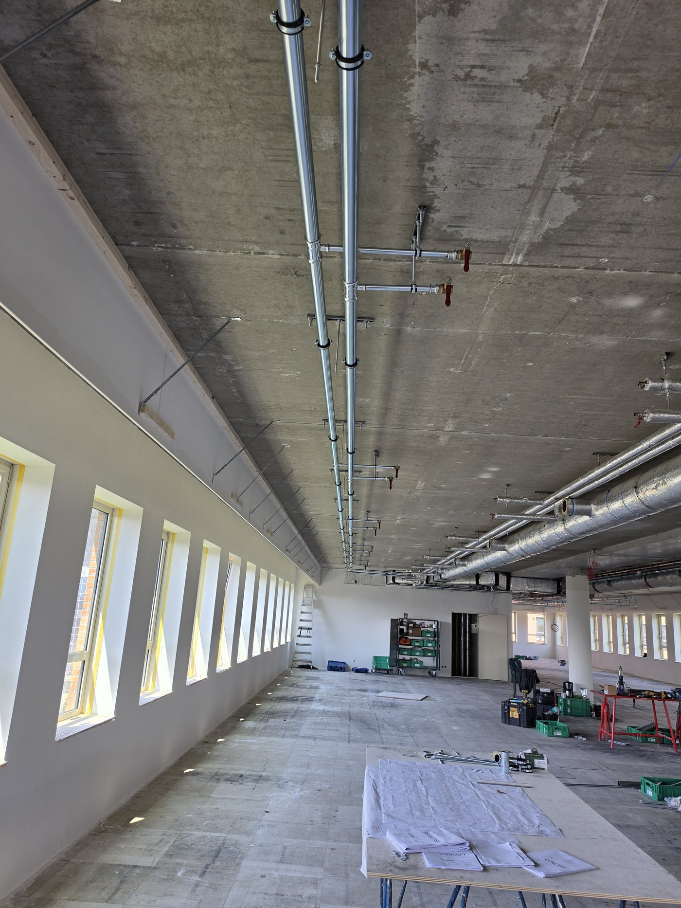 Installatiewerk langs het plafond van een kantoorverdieping in aanbouw, met een rij ramen op de achtergrond